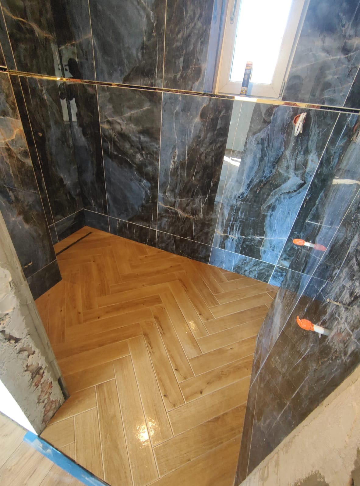 Ukończona łazienka z ciemnymi marmurkowymi płytkami ściennymi i podłogą z płytek imitujących drewno ułożonych w jodełkę, widoczne niedokończone wykończenia przy wejściu.