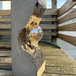 Betonowa lampa z żarówką Edison, wsparta na pręcie zbrojeniowym, ustawiona na drewnianym tarasie w słoneczny dzień.