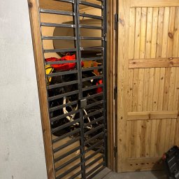 PMD-Design Sp. z o.o. - Metalowa krata z poziomymi prętami, w drewnianej futrynie, zasłaniająca widok na garaż z rowerami. Surowe wykończenie, widoczne detale spawów.