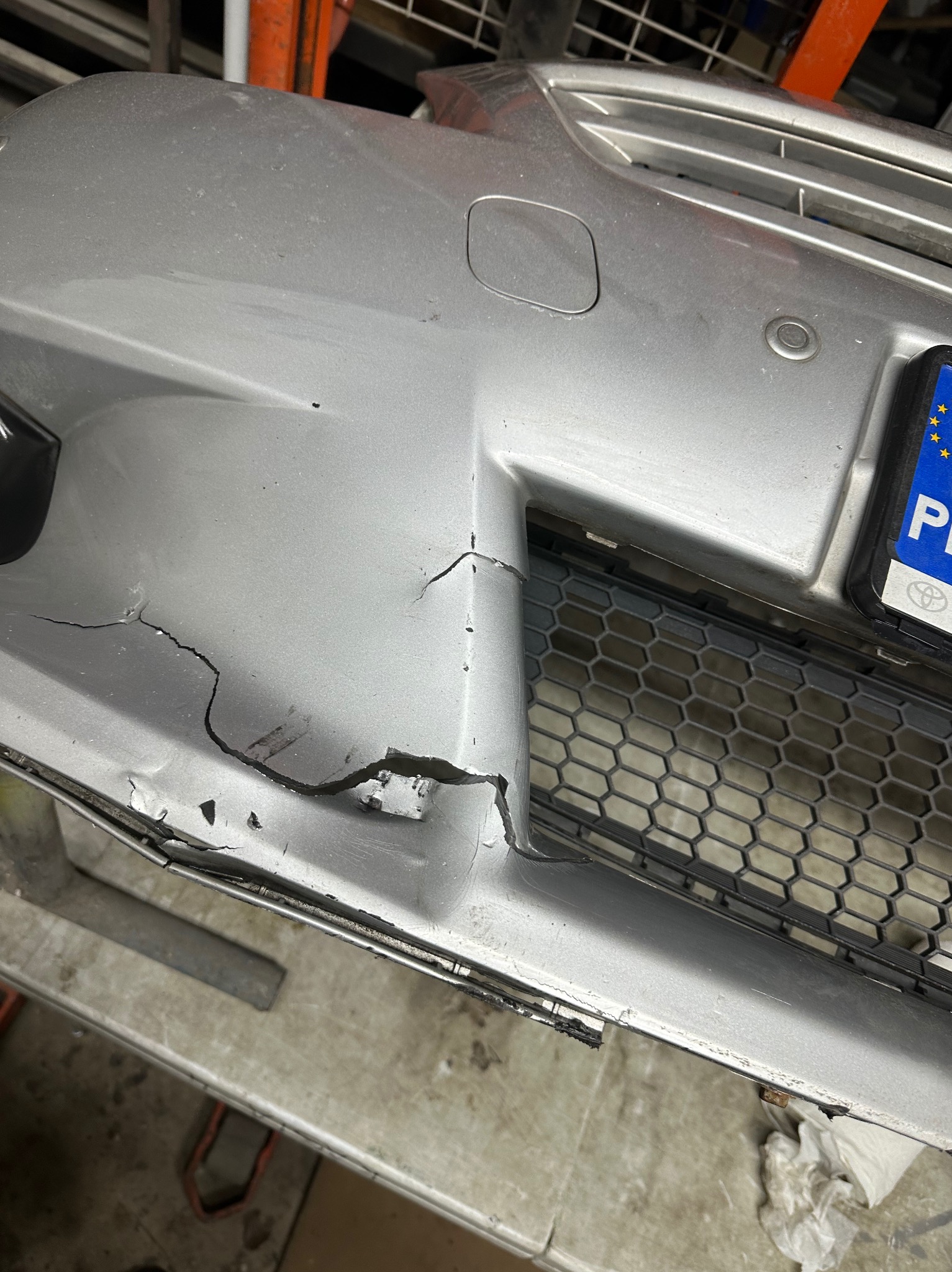 Uszkodzony, srebrny zderzak samochodowy z widocznymi pęknięciami i ubytkami lakieru w warsztacie.