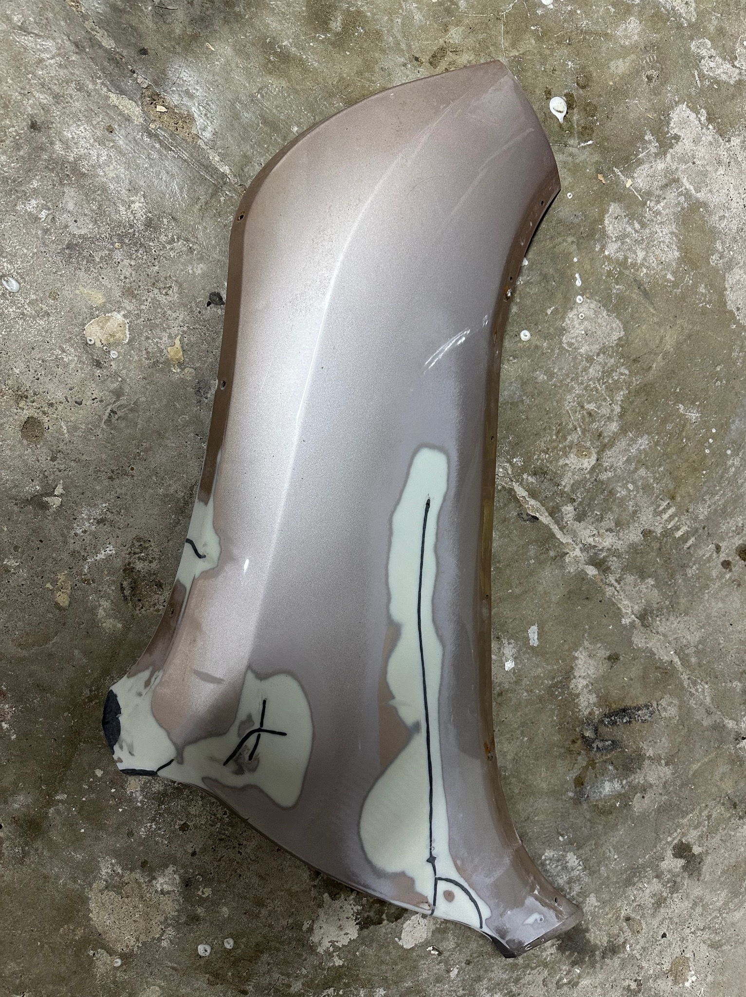 Element karoserii samochodowej w trakcie naprawy blacharskiej, z widocznymi szpachlami i zaznaczeniami, leżący na betonowej posadzce.