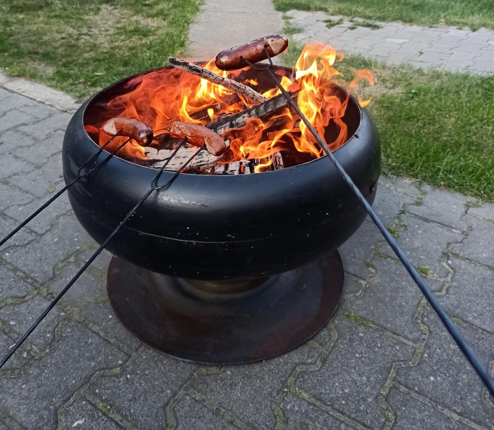 Ognisko w metalowym palenisku z trzema kiełbaskami nad płomieniami, wbitymi na długie, metalowe szpikulce, na tle kostki brukowej i trawy.