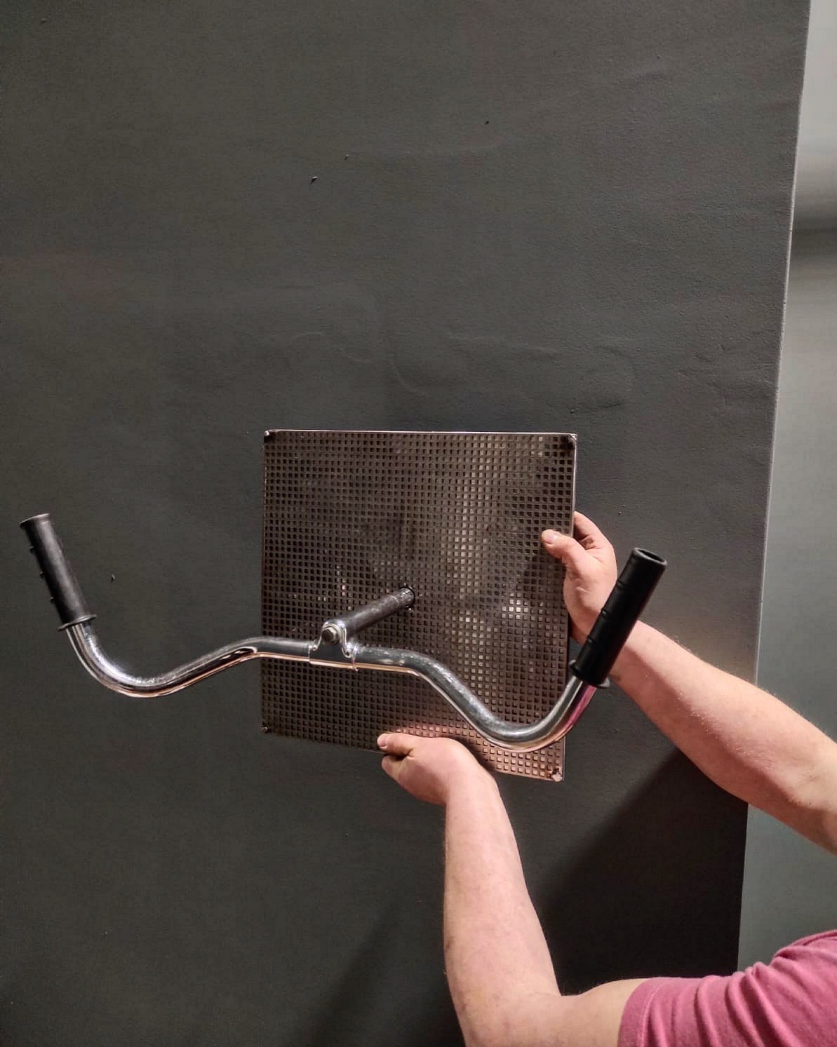 Mężczyzna w Swarzędzu prezentuje metalowy uchwyt z kierownicą rowerową przymocowaną do perforowanej blachy na tle szarej ściany.