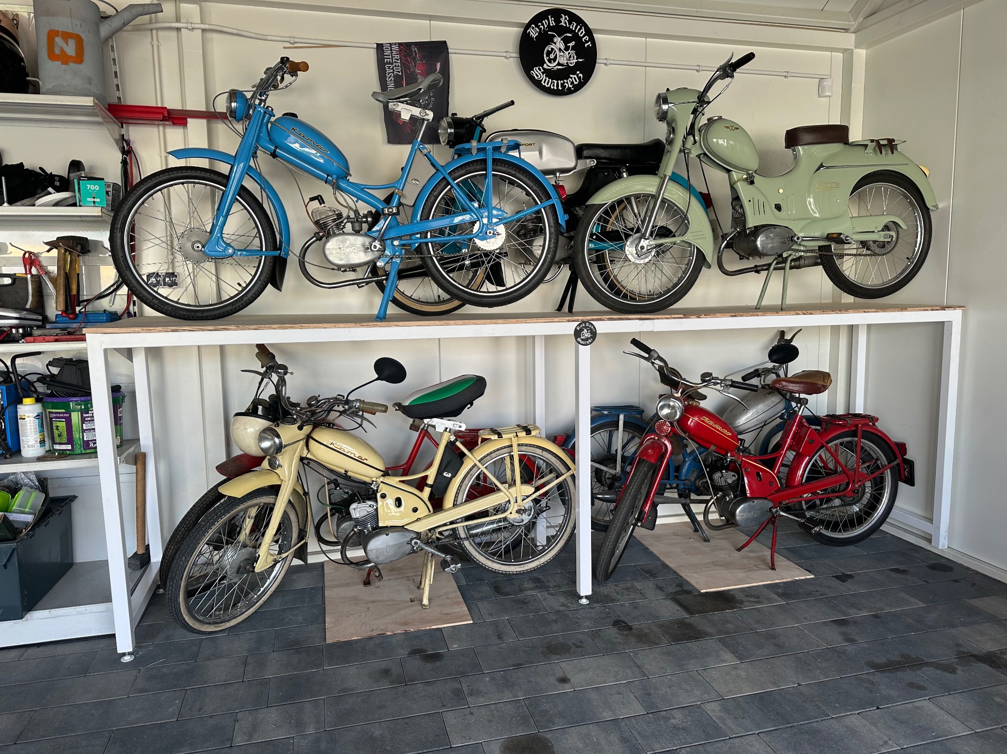Kolekcja sześciu odrestaurowanych, zabytkowych motorowerów Komar w różnych kolorach, ustawionych na białych półkach w garażu z szarymi płytkami podłogowymi.