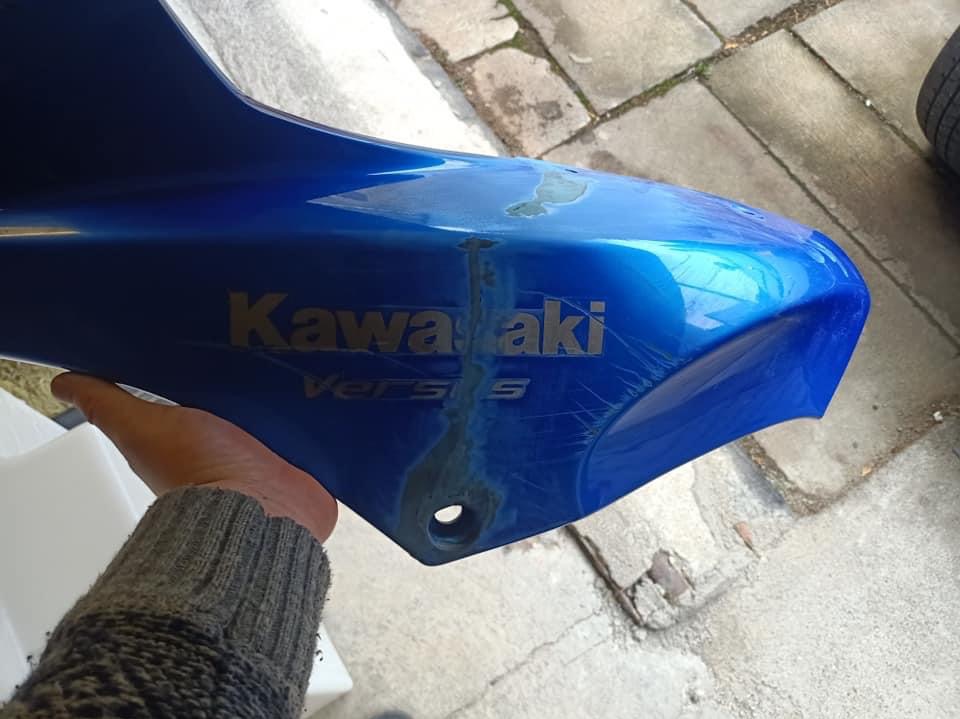 Uszkodzony, niebieski element karoserii motocykla Kawasaki Versys, z widocznymi rysami i śladami naprawy, trzymany w dłoni na tle betonowej nawierzchni.