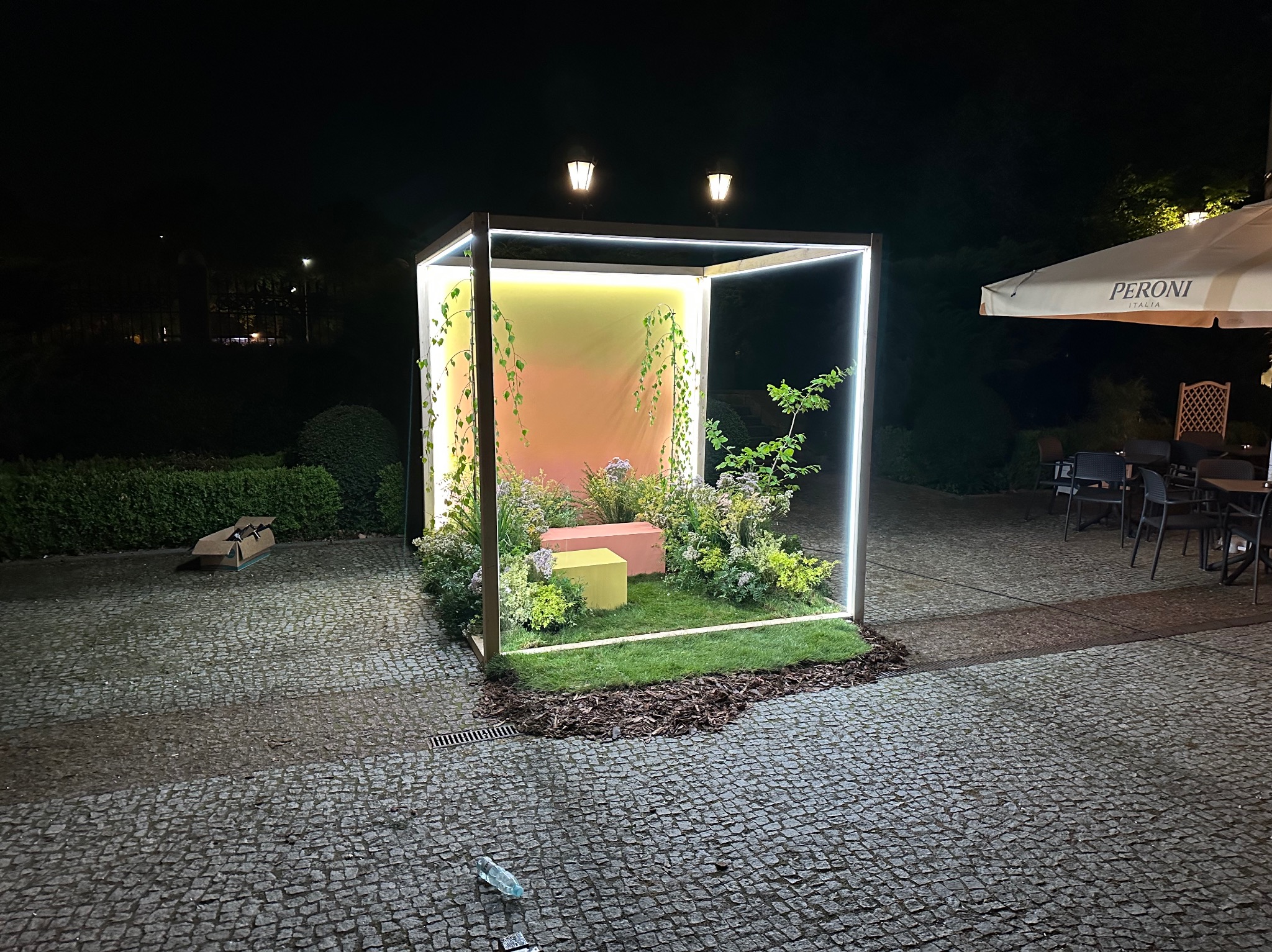 Oświetlona, metalowa konstrukcja sześcienna z roślinami i dekoracyjnymi elementami wewnątrz, ustawiona na brukowanym placu nocą, z parasolem Peroni w tle.