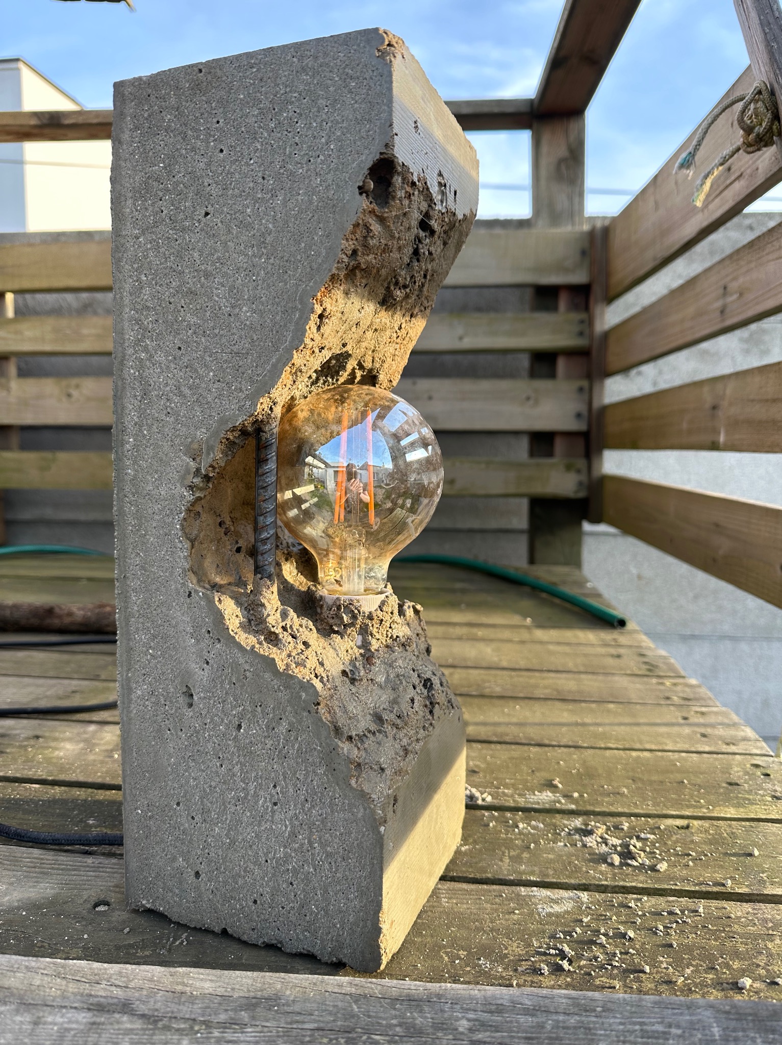 Betonowa lampa z żarówką Edison, wsparta na pręcie zbrojeniowym, ustawiona na drewnianym tarasie w słoneczny dzień.