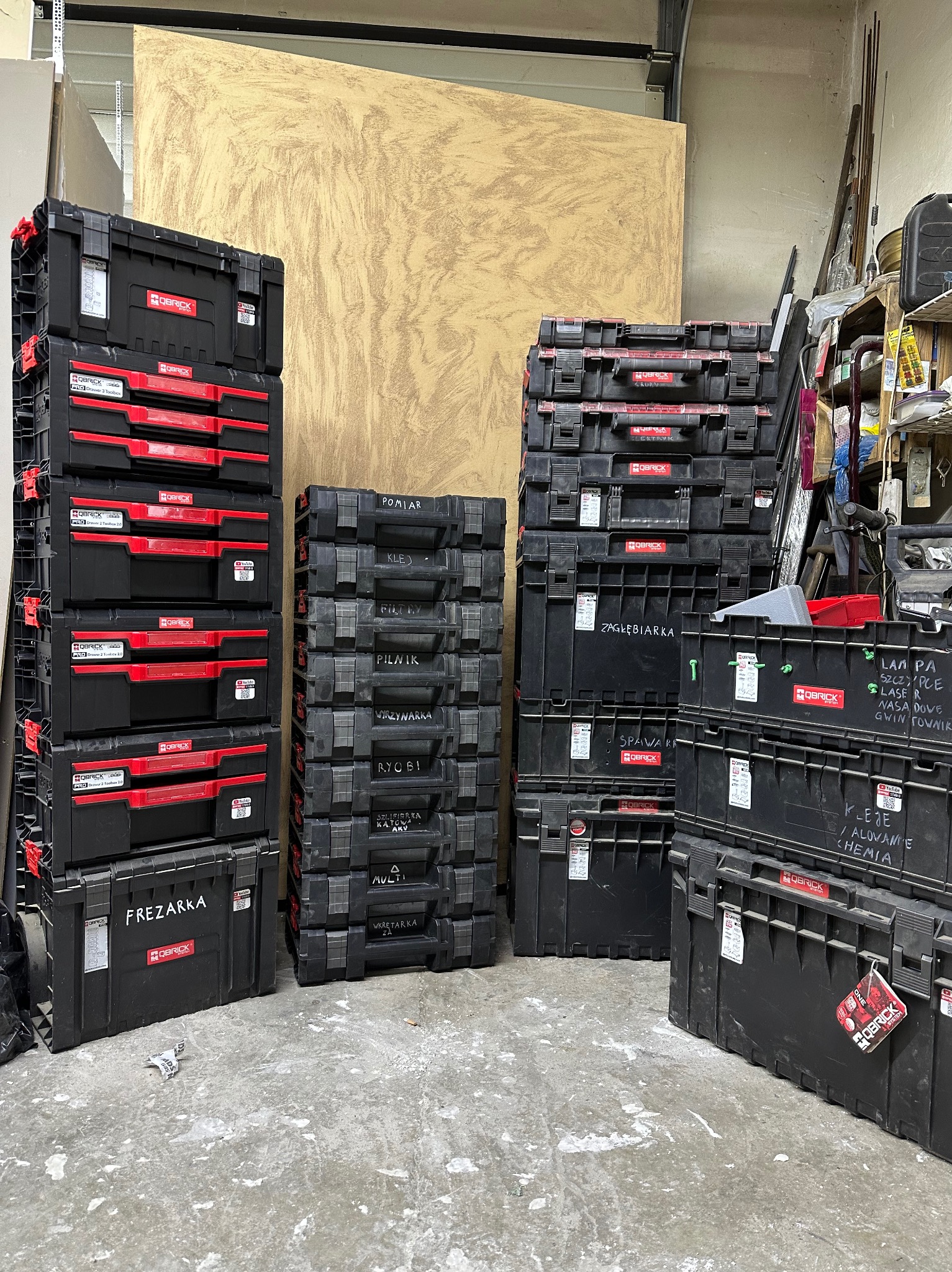 Stosy czarnych skrzynek narzędziowych QBRICK, niektóre z czerwonymi szufladami, ustawione w warsztacie; na niektórych pudełkach widoczne napisy odręczne, np. 'Frezarka', 'Spawark', 'Zagłębiarka'...