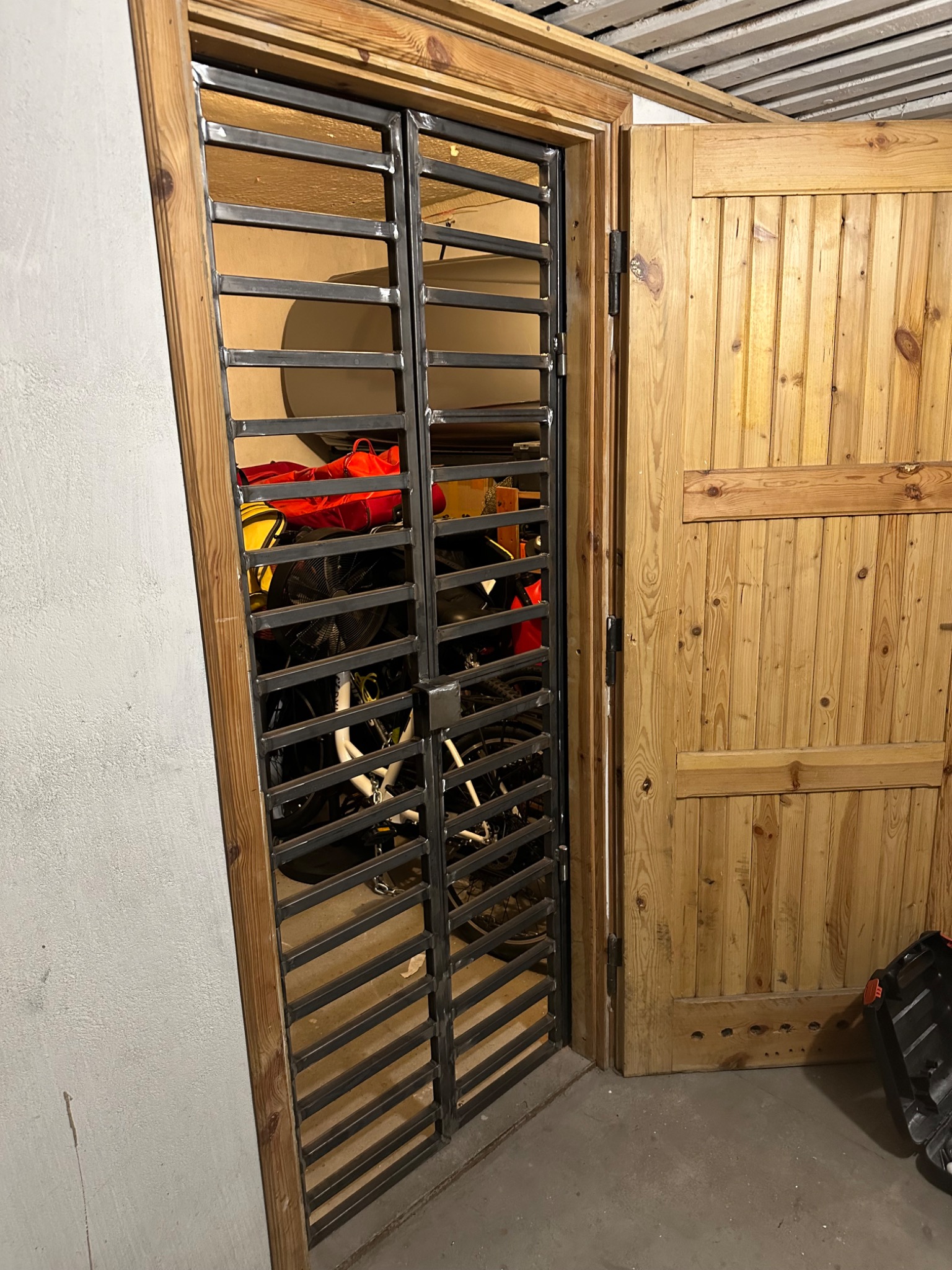 Metalowa krata z poziomymi prętami, w drewnianej futrynie, zasłaniająca widok na garaż z rowerami. Surowe wykończenie, widoczne detale spawów.