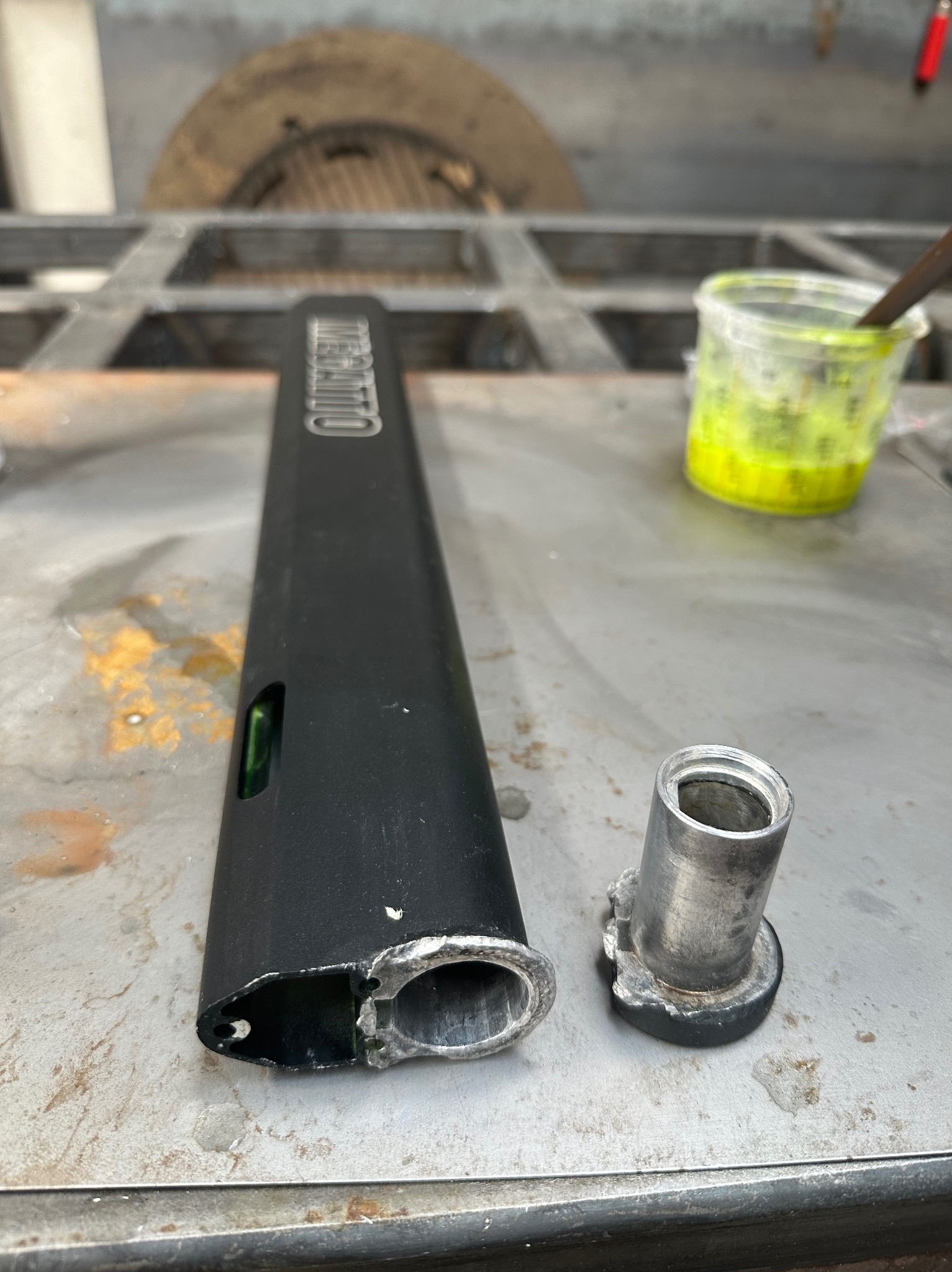 Element metalowy z widocznymi śladami obróbki, prawdopodobnie spawania, leży na metalowym stole warsztatowym obok pojemnika z płynem.