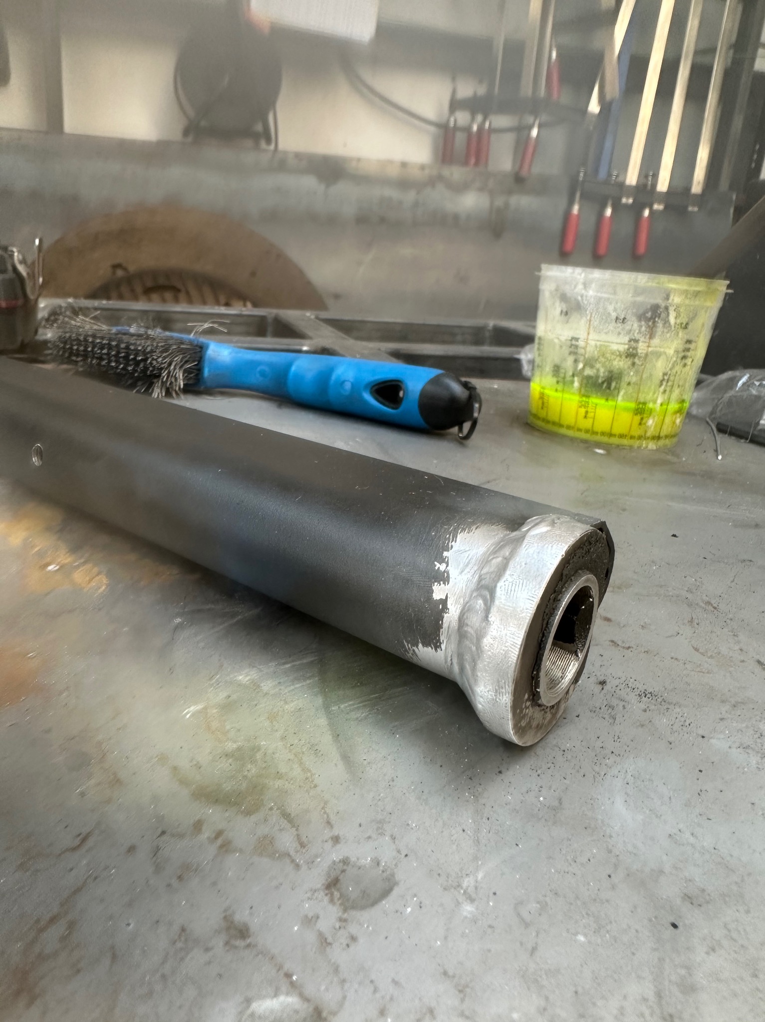 Czarny metalowy profil z widocznym spawem na końcu, leżący na metalowym stole warsztatowym. Obok szczotka druciana i pojemnik z płynem.