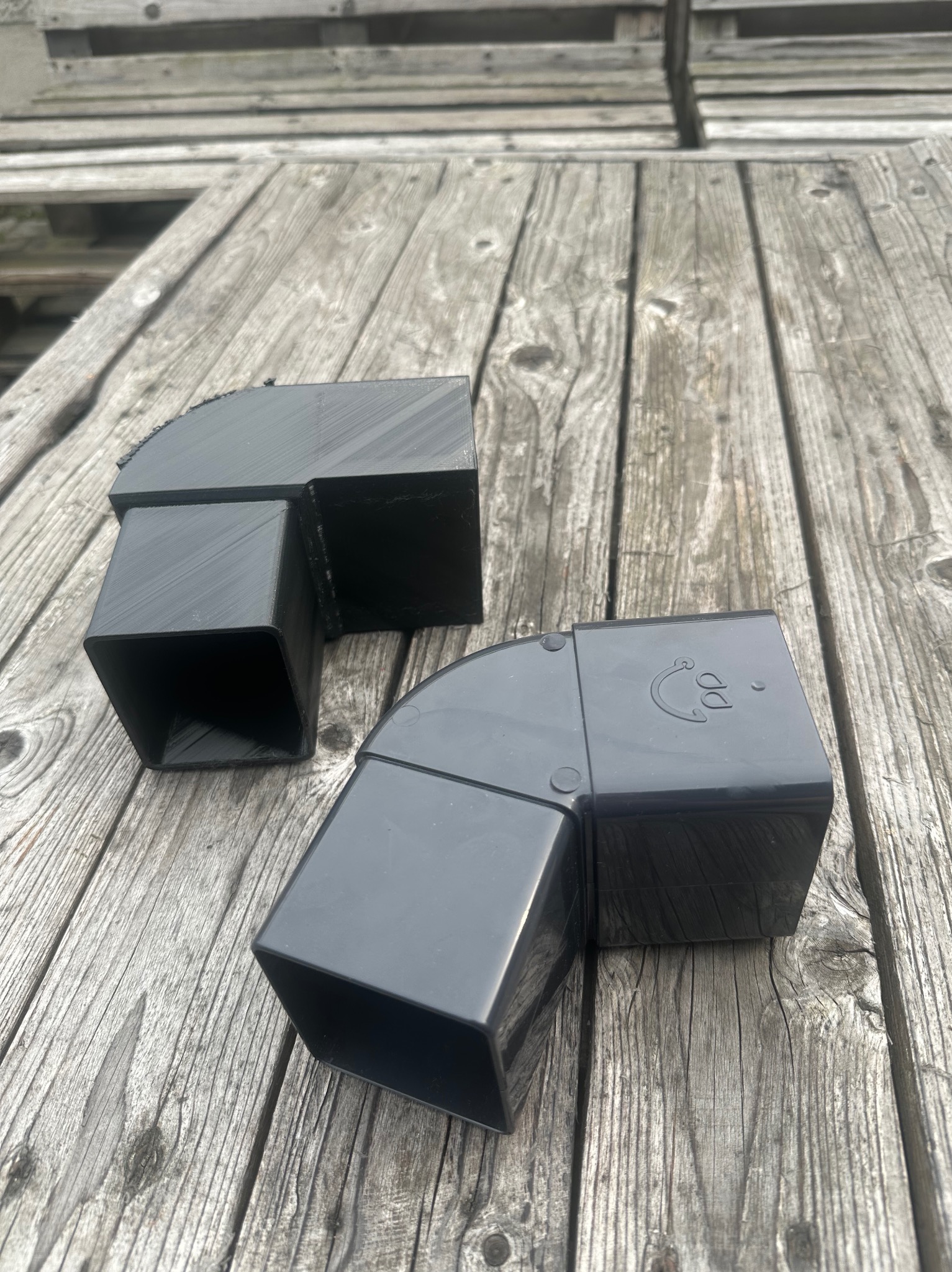 Dwa czarne, kwadratowe łączniki rurowe z tworzywa na szarym, drewnianym stole. Jeden z nadrukiem. Widoczne ślady druku 3D.