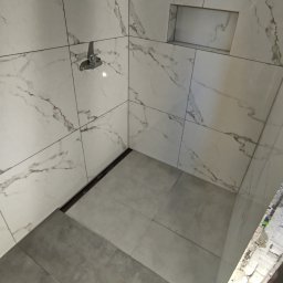 Karol Kazaniecki - Wykończona kabina prysznicowa z marmurowymi płytkami, wbudowaną półką i odpływem liniowym, widoczne niedokończone wykończenie ściany po prawej stronie.