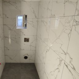 Karol Kazaniecki - Wnętrze łazienki w trakcie remontu, widoczne białe płytki imitujące marmur na ścianach, szara podłoga, okno z otwartym skrzydłem, wystające rury i puszki elektryczne przed montażem osprzętu.