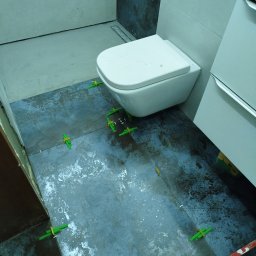 Karol Kazaniecki - Montaż płytek podłogowych w łazience, widoczne krzyżyki dystansowe, toaleta podwieszana i umywalka z szafką.