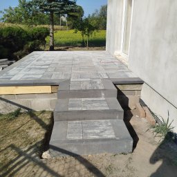 Karol Kazaniecki - Wykończony taras z szarej kostki brukowej z trzema stopniami prowadzącymi do budynku o surowej, jasnoszarej elewacji. Część tarasu jeszcze w trakcie prac, widoczne elementy drewniane. W tle zieleń...