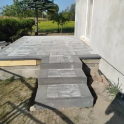 Wykończony taras z szarej kostki brukowej z trzema stopniami prowadzącymi do budynku o surowej, jasnoszarej elewacji. Część tarasu jeszcze w trakcie prac, widoczne elementy drewniane. W tle zieleń...