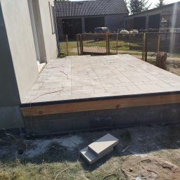 Karol Kazaniecki - Wykończony taras z jasnej kostki brukowej przylegający do budynku, z widocznym podmurowaniem z bloczków betonowych i drewnianą konstrukcją nośną oraz niezabudowanymi elementami kostki obok.