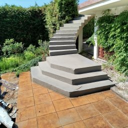 Karol Kazaniecki - Betonowe schody zewnętrzne prowadzące do budynku, otoczone zielenią, z fragmentem zdemontowanej blachy na pierwszym planie i brązowymi płytkami chodnikowymi.