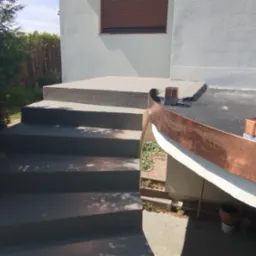 Zewnętrzne schody betonowe prowadzące do budynku z obróbkami blacharskimi z miedzi, widoczne ślady po niedawnym malowaniu betonu.