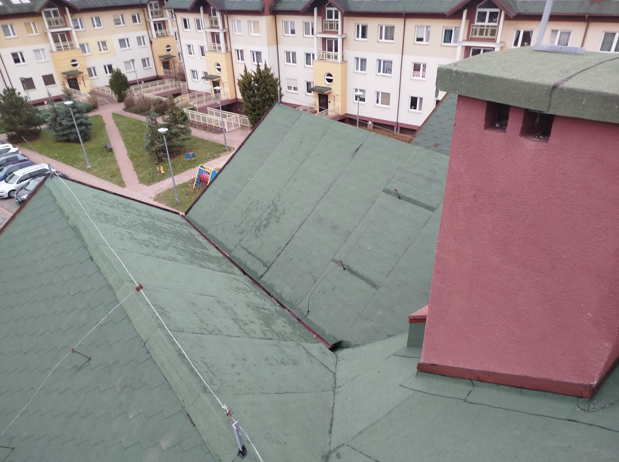 Zielony dach z papy termozgrzewalnej i czerwony komin na tle zabudowy wielorodzinnej z zielonym dziedzińcem.