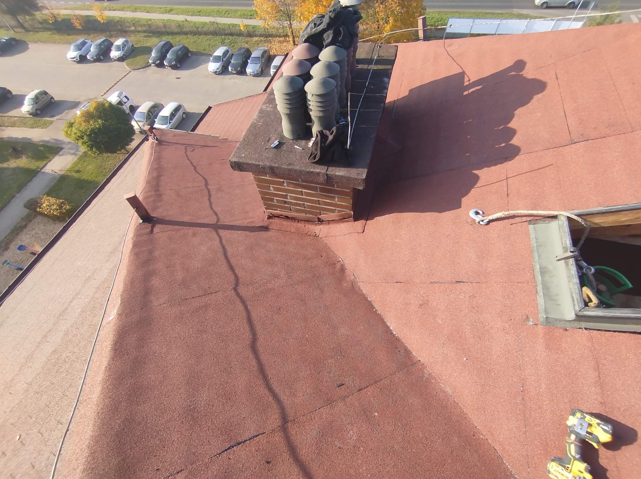 Czerwony dach pokryty papą z kominem i wentylacją, widoczne narzędzia i lina zabezpieczająca, perspektywa z góry.
