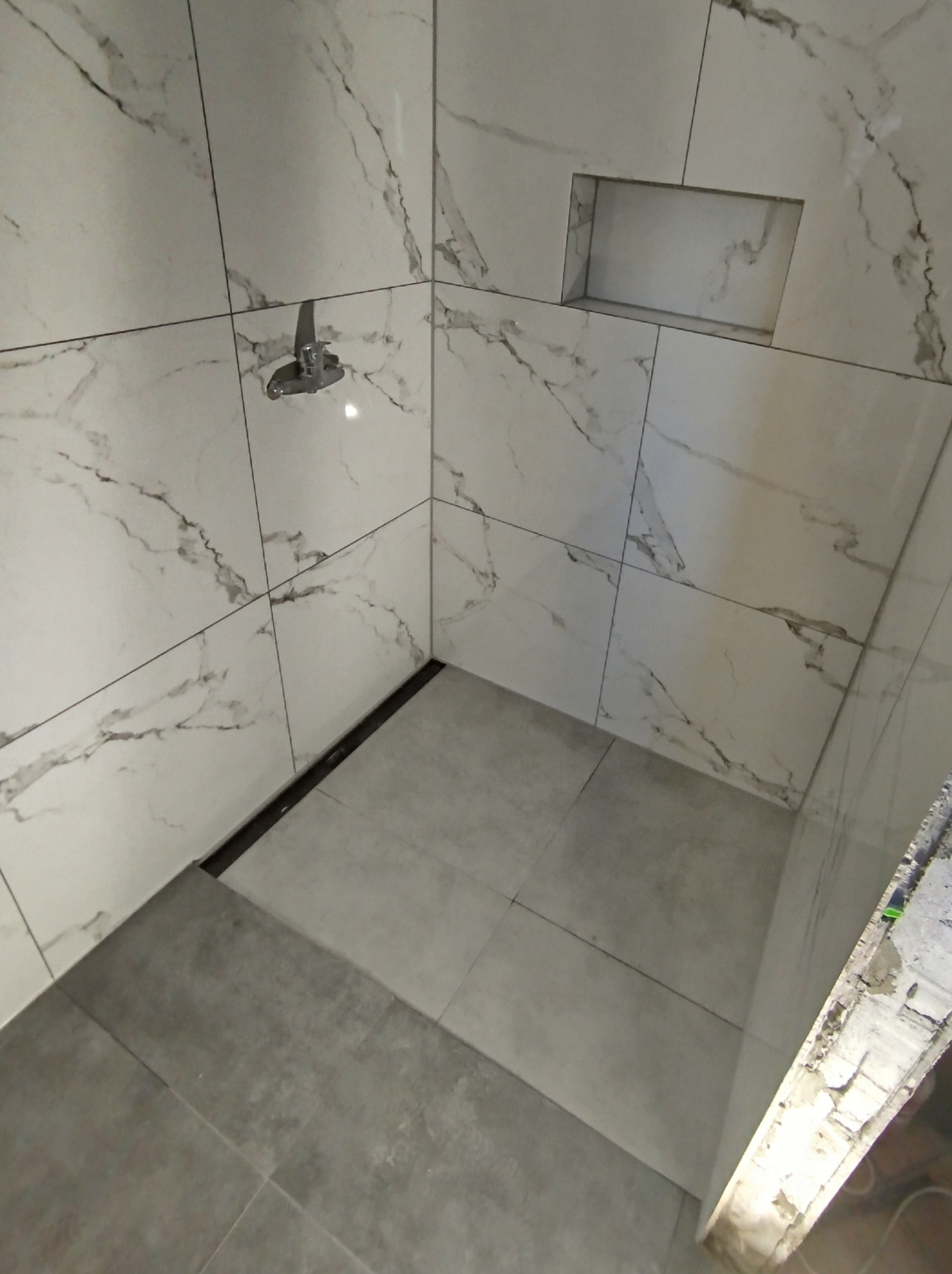 Wykończona kabina prysznicowa z marmurowymi płytkami, wbudowaną półką i odpływem liniowym, widoczne niedokończone wykończenie ściany po prawej stronie.