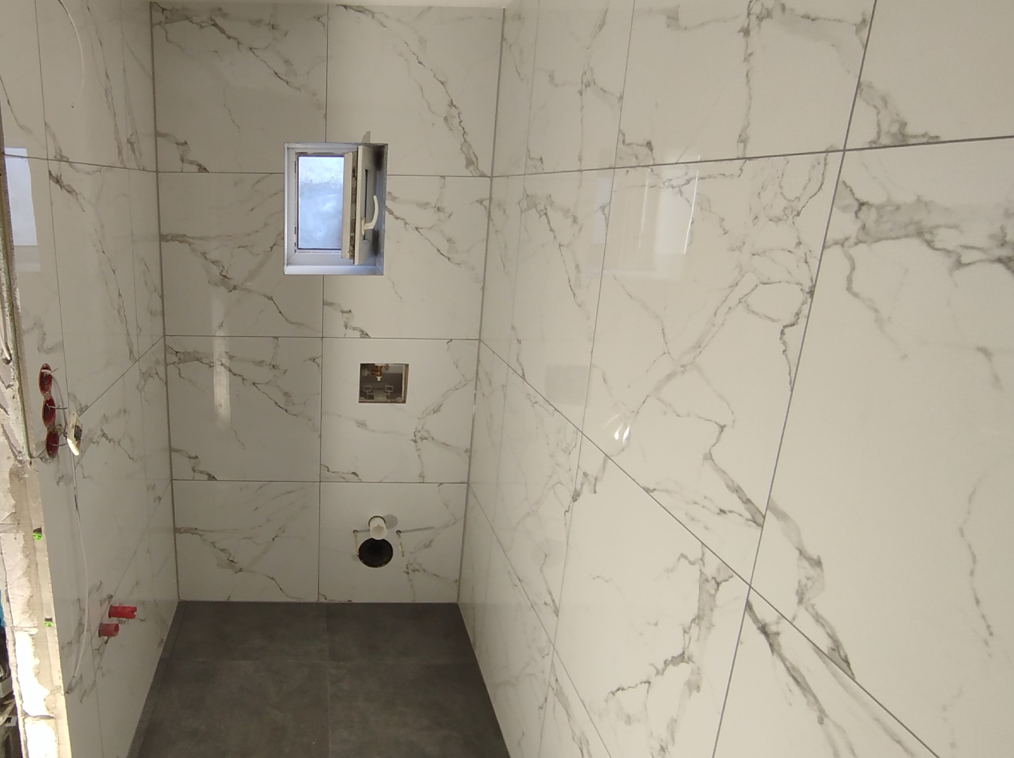 Wnętrze łazienki w trakcie remontu, widoczne białe płytki imitujące marmur na ścianach, szara podłoga, okno z otwartym skrzydłem, wystające rury i puszki elektryczne przed montażem osprzętu.