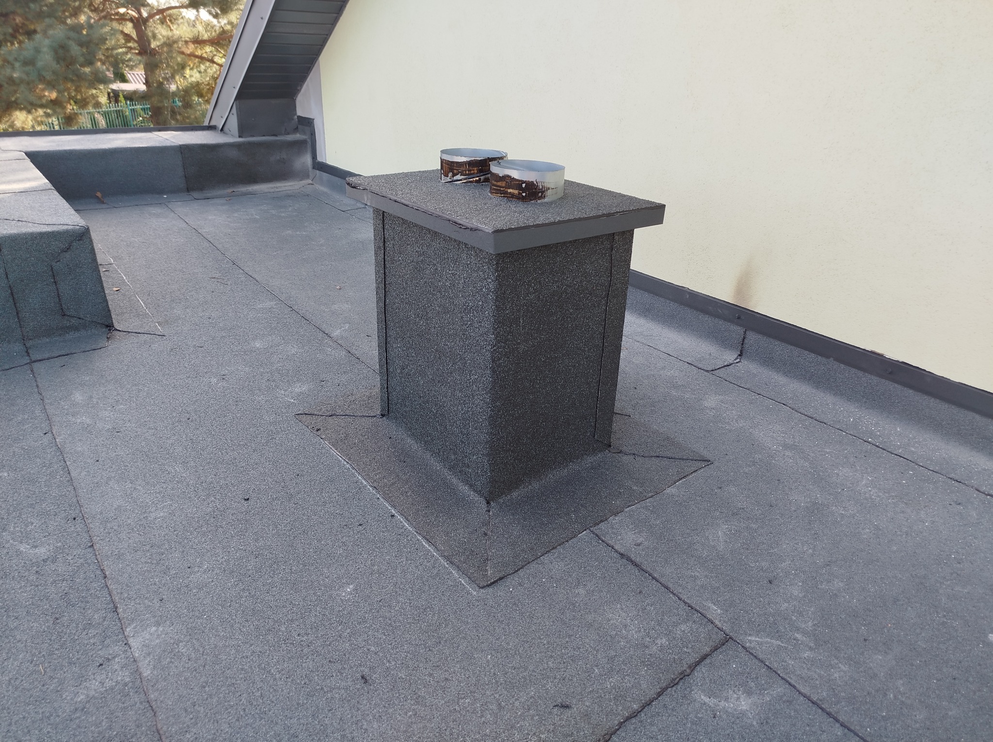 Wykończony komin z obróbkami blacharskimi na dachu pokrytym papą w Ciechocinie, widoczne dwa metalowe elementy na górze komina.