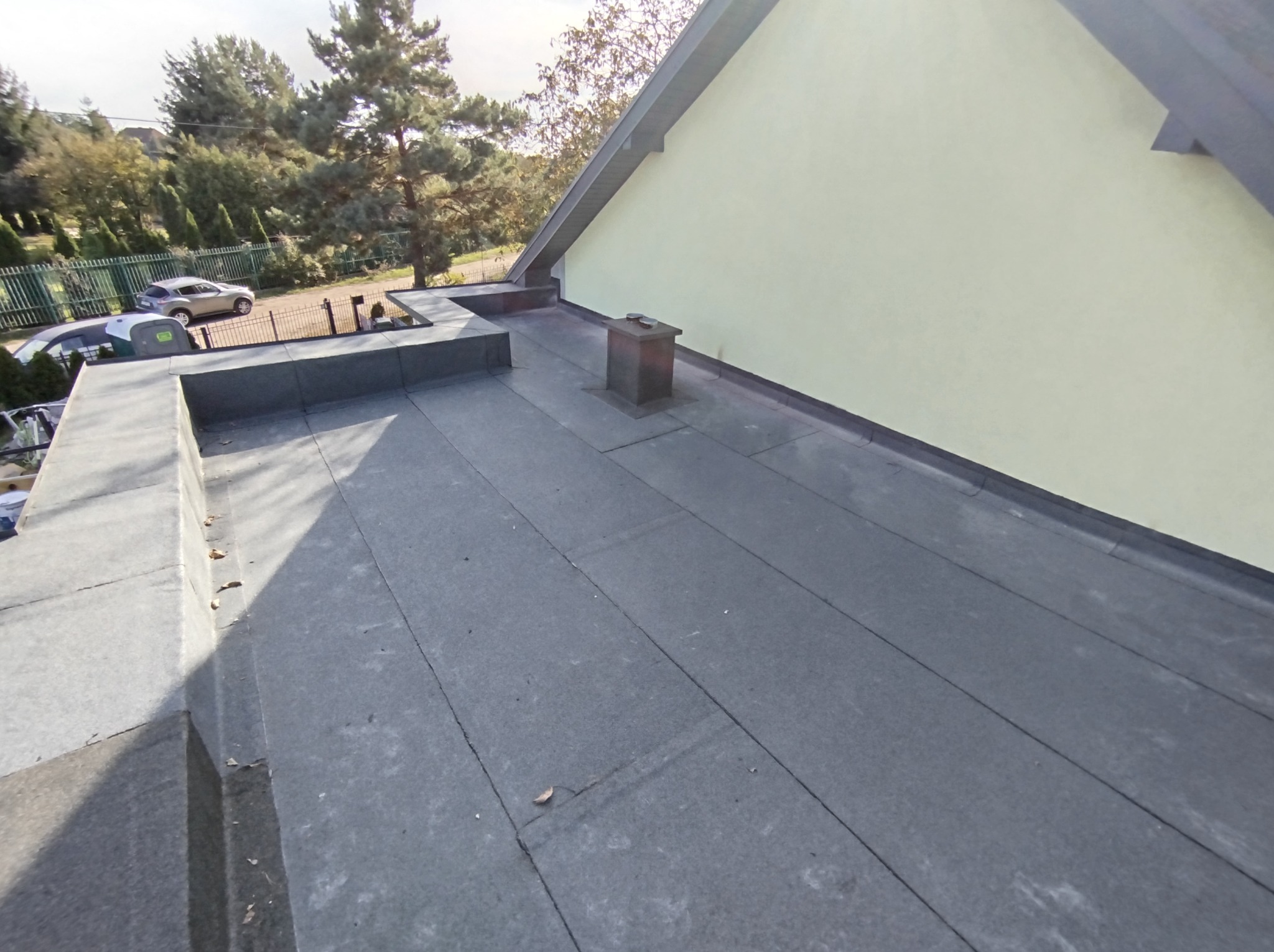 Widok na płaski dach pokryty papą, z kominem i obróbkami blacharskimi, na tle jasnej ściany budynku i zieleni drzew.