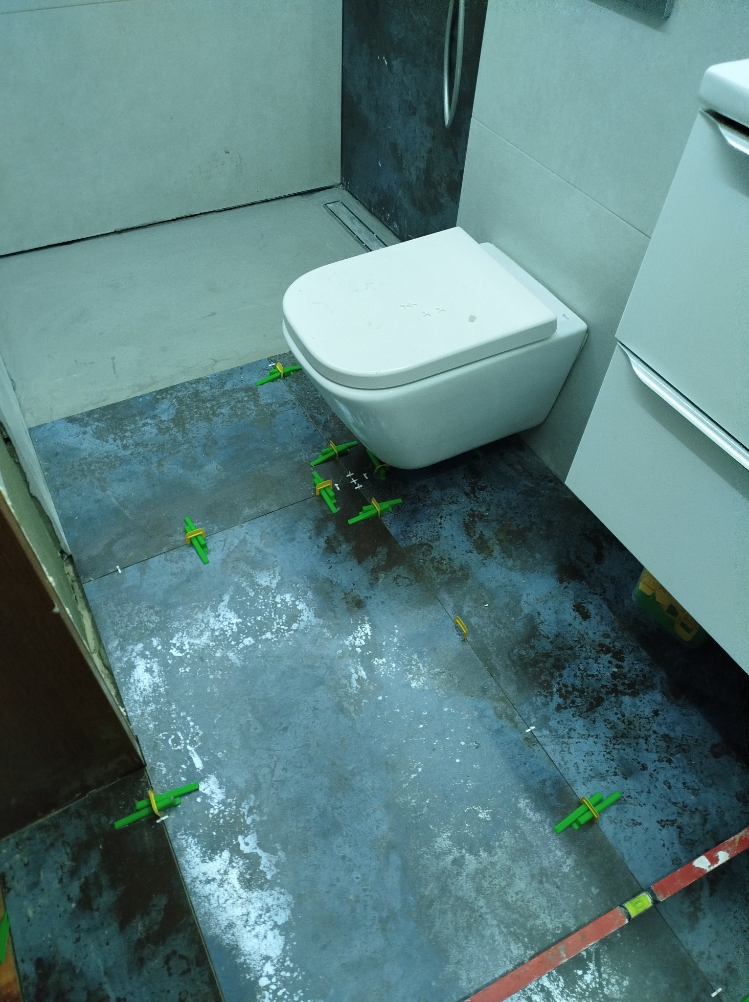 Montaż płytek podłogowych w łazience, widoczne krzyżyki dystansowe, toaleta podwieszana i umywalka z szafką.