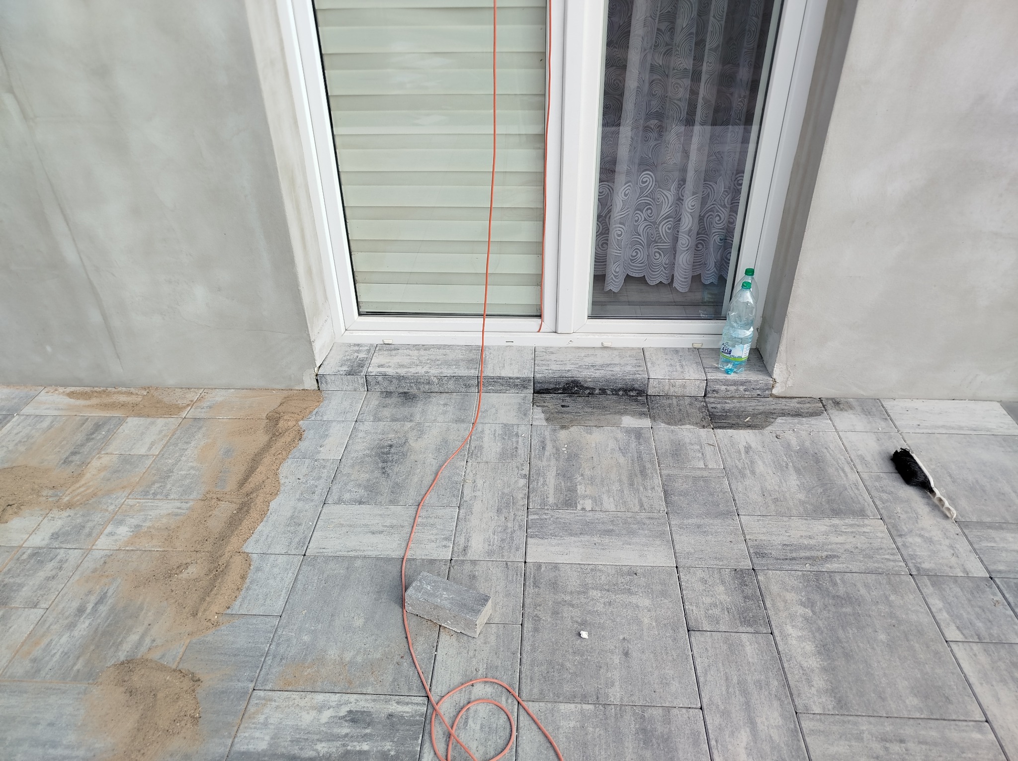 Przygotowanie podłoża pod okno z nową elewacją: ułożona kostka brukowa, piasek do wyrównania, przewód zasilający i butelka wody.