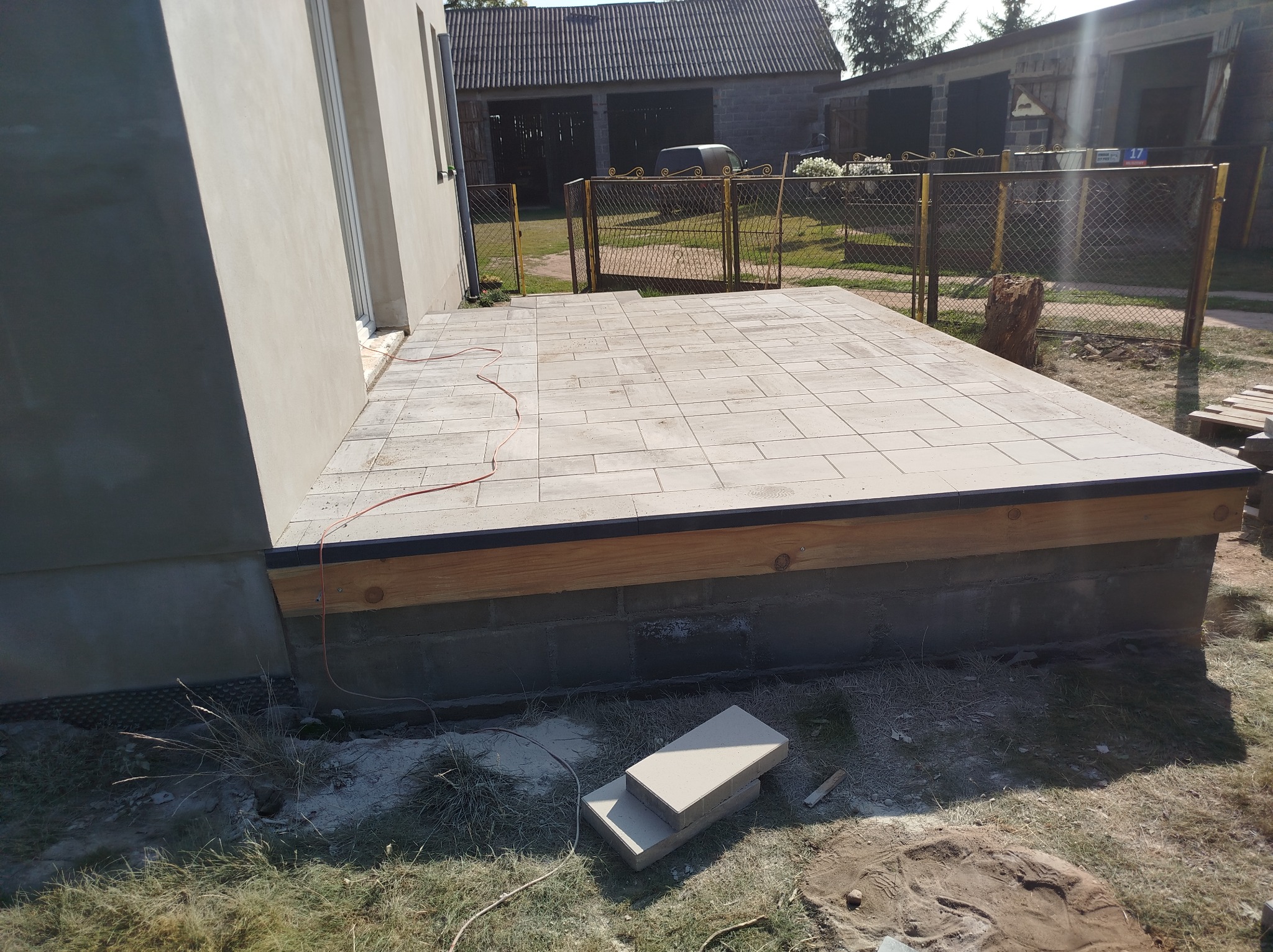 Wykończony taras z jasnej kostki brukowej przylegający do budynku, z widocznym podmurowaniem z bloczków betonowych i drewnianą konstrukcją nośną oraz niezabudowanymi elementami kostki obok.