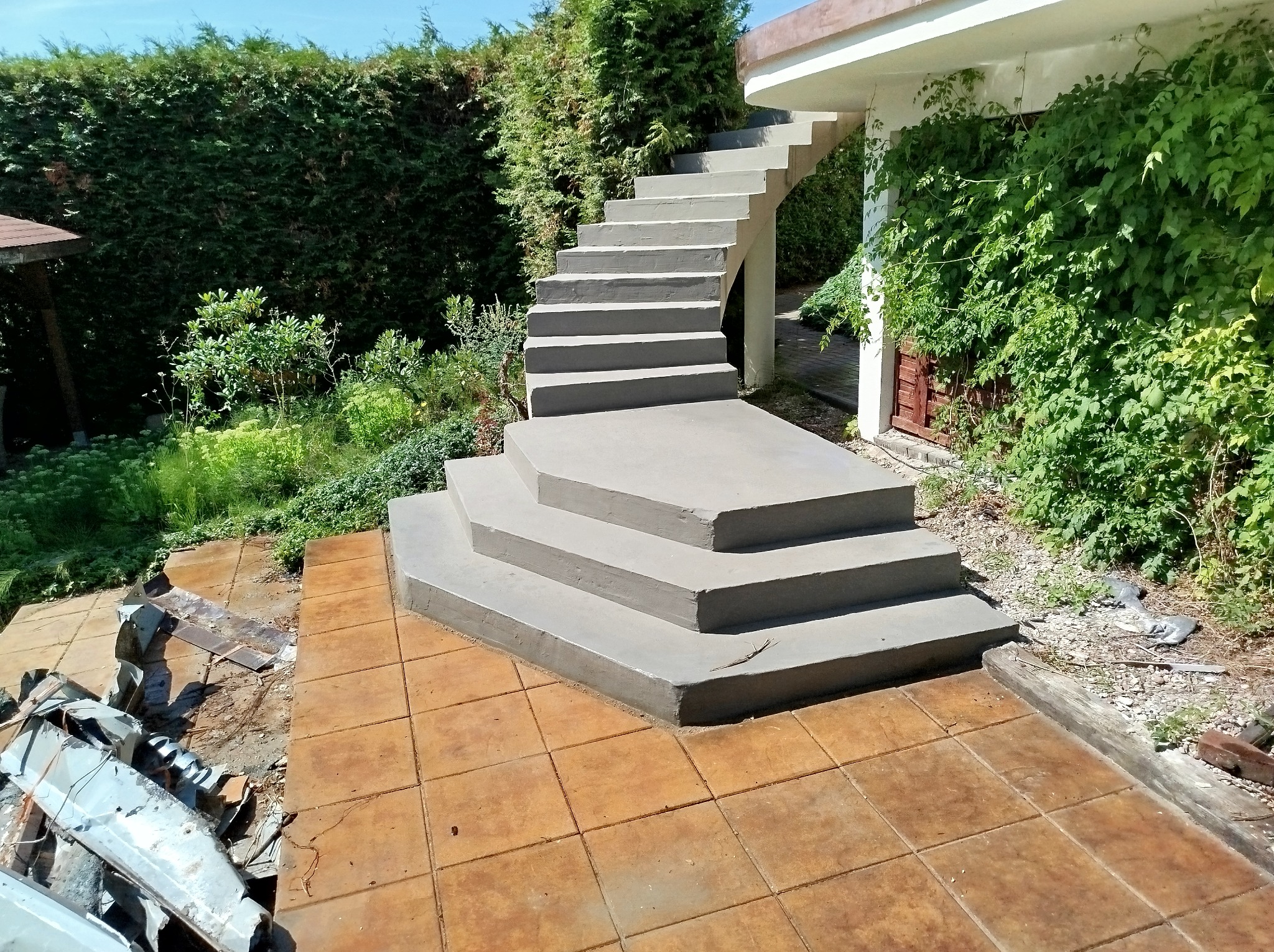 Betonowe schody zewnętrzne prowadzące do budynku, otoczone zielenią, z fragmentem zdemontowanej blachy na pierwszym planie i brązowymi płytkami chodnikowymi.