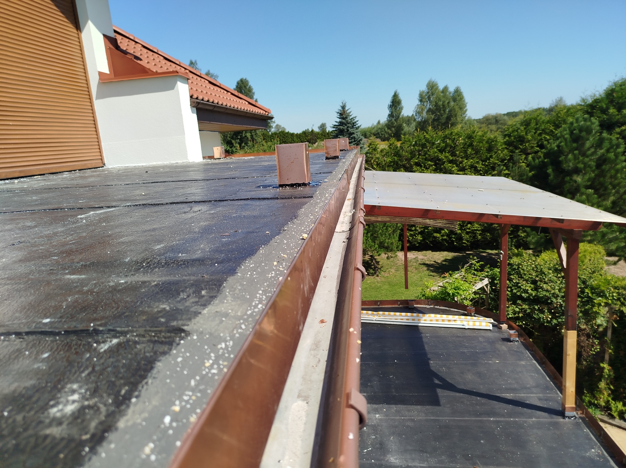 Widok z góry na fragment dachu z brązowymi obróbkami blacharskimi i rynną, w tle zieleń drzew i fragment budynku z roletą zewnętrzną.