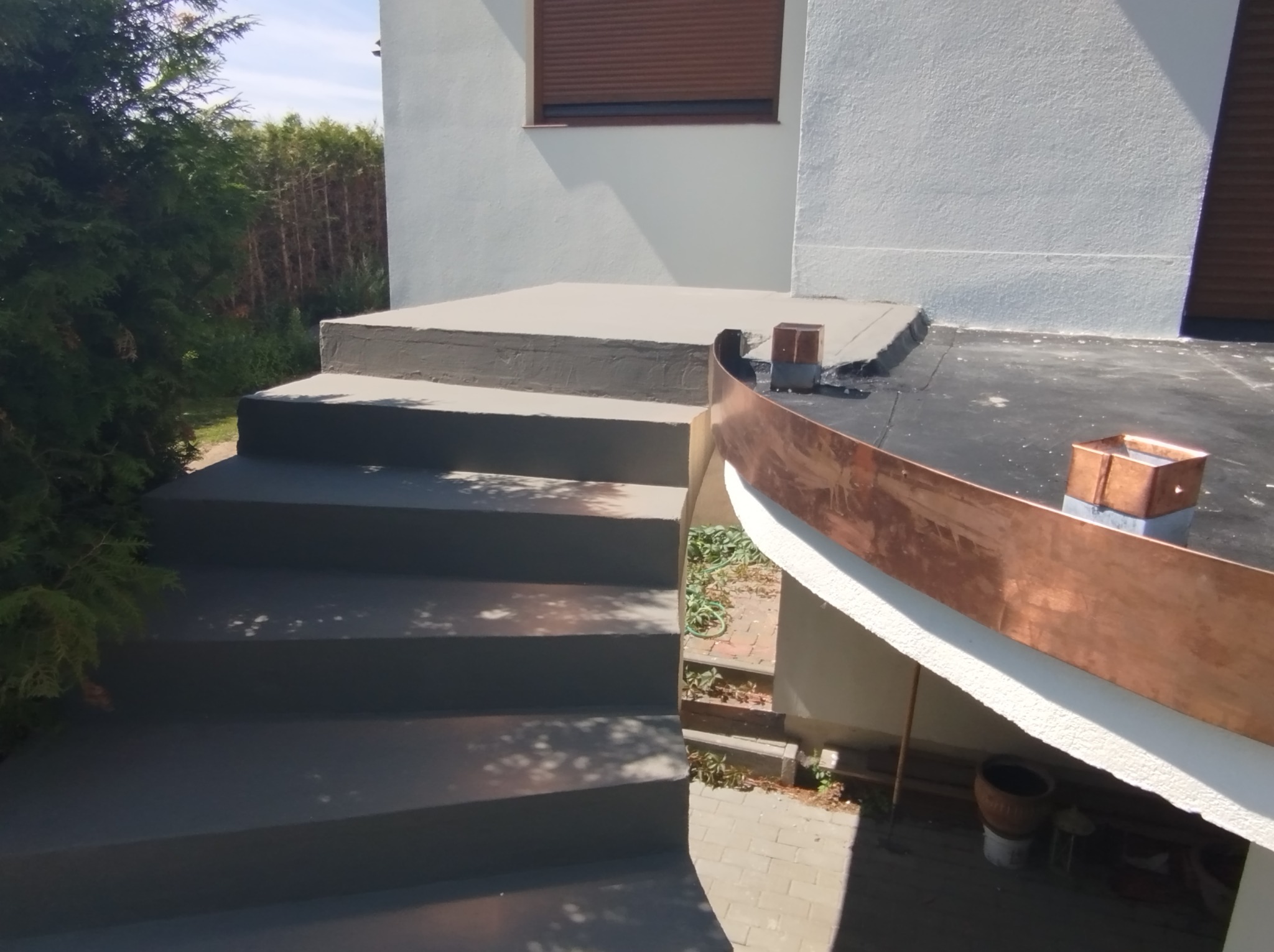 Zewnętrzne schody betonowe prowadzące do budynku z obróbkami blacharskimi z miedzi, widoczne ślady po niedawnym malowaniu betonu.