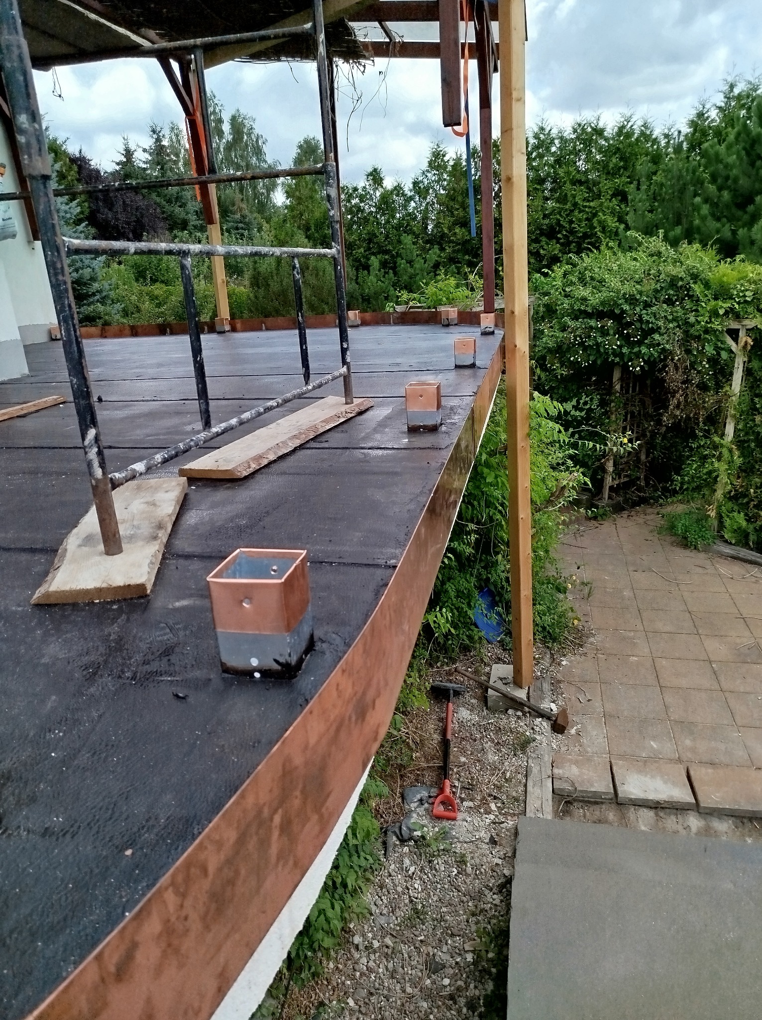 Montaż miedzianych obróbek blacharskich na dachu tarasu z widocznym rusztowaniem, elementami konstrukcyjnymi i narzędziami w tle bujna roślinność.