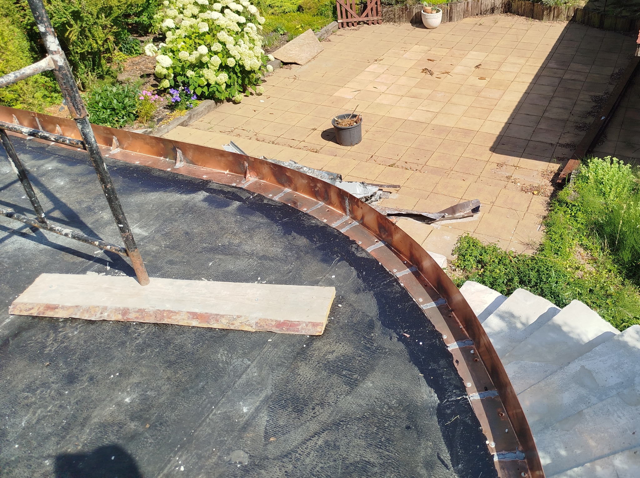 Montaż miedzianej obróbki blacharskiej na okrągłym dachu, widok z góry, drabina oparta o dach, w tle patio z płytkami i roślinność.