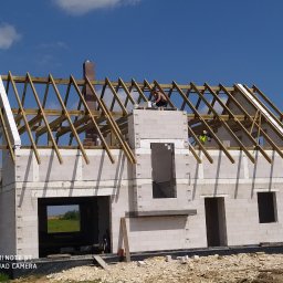 FU Lukas Łukasz Luboń - Wznoszenie ścian domu z betonu komórkowego Ytong, widoczne drewniane elementy konstrukcji dachu i komin z cegły, pracownik na dachu w trakcie murowania komina.