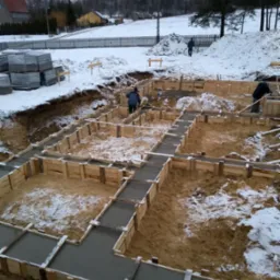 Zimowy krajobraz budowy fundamentów: drewniane szalunki wypełnione świeżym betonem, częściowo pokryte śniegiem, widoczni pracownicy w oddali, w tle ośnieżone domy i drzewa.