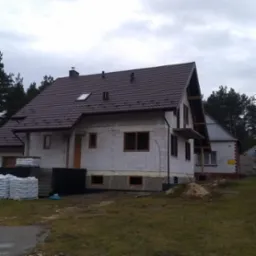 Niedokończony dom z białych bloczków z brązowym dachem, widoczne okna bez szyb i palety z materiałami budowlanymi na podwórku, w tle drzewa i pochmurne niebo.