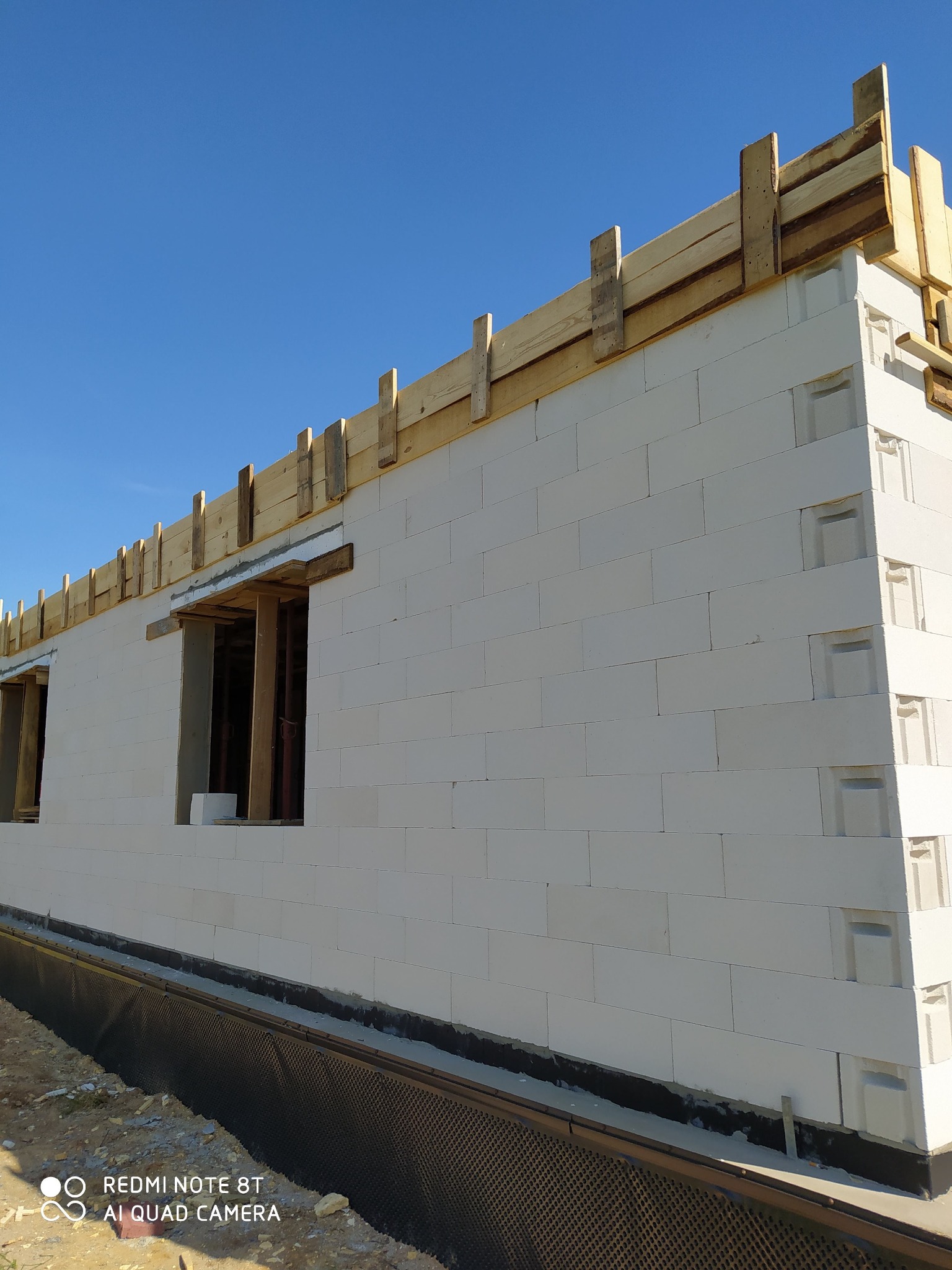 Wznoszenie ścian domu z białych bloczków, widoczne drewniane szalunki nad oknami i na górze muru, zabezpieczenie fundamentów folią kubełkową.