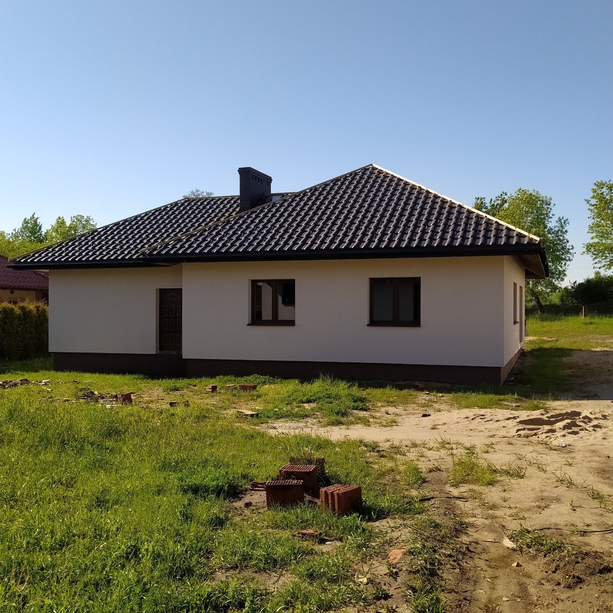 Niedawno wybudowany, parterowy dom murowany z ciemną dachówką, widocznymi cegłami na trawie i świeżo posadzoną trawą.