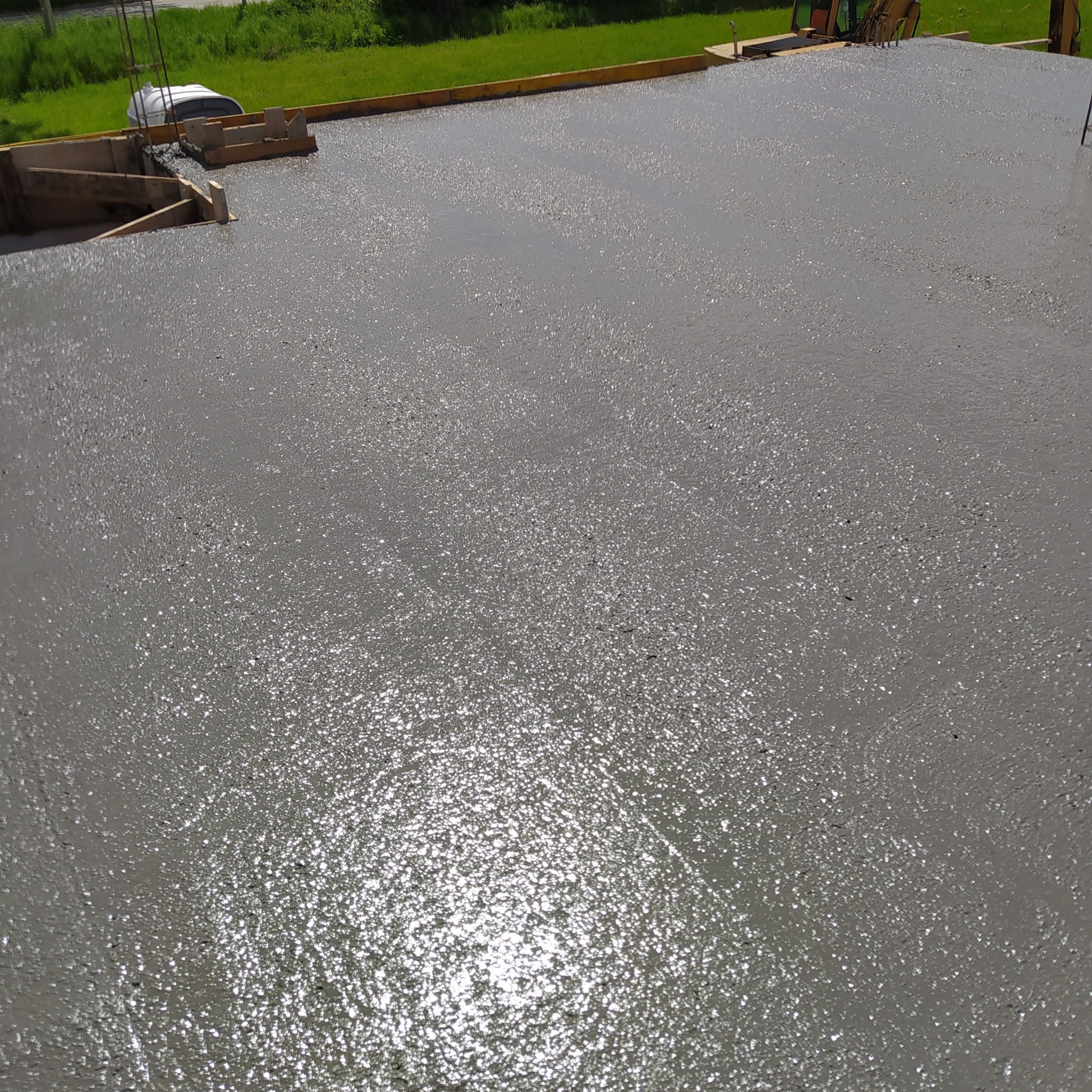 Świeżo wylany, mokry beton fundamentowy z widocznym szalunkiem drewnianym i zbrojeniem, w tle zielona trawa i fragment koparki.