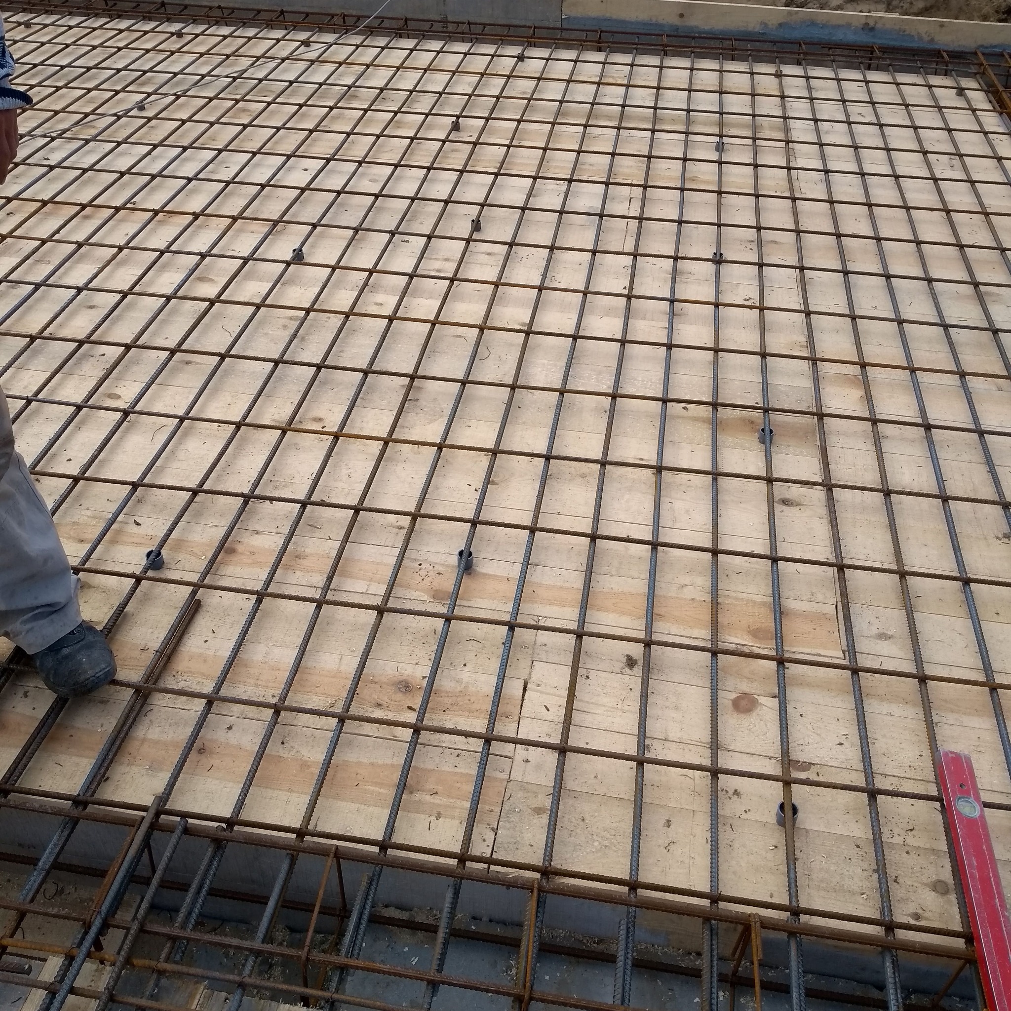 Zbrojenie fundamentu na drewnianym szalunku, widoczne dystanse z tworzywa, fragment stopy robotnika i poziomica.