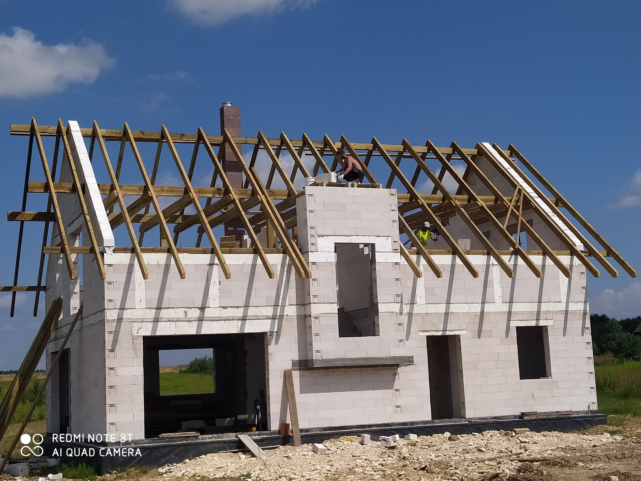 Wznoszenie ścian domu z betonu komórkowego Ytong, widoczne drewniane elementy konstrukcji dachu i komin z cegły, pracownik na dachu w trakcie murowania komina.
