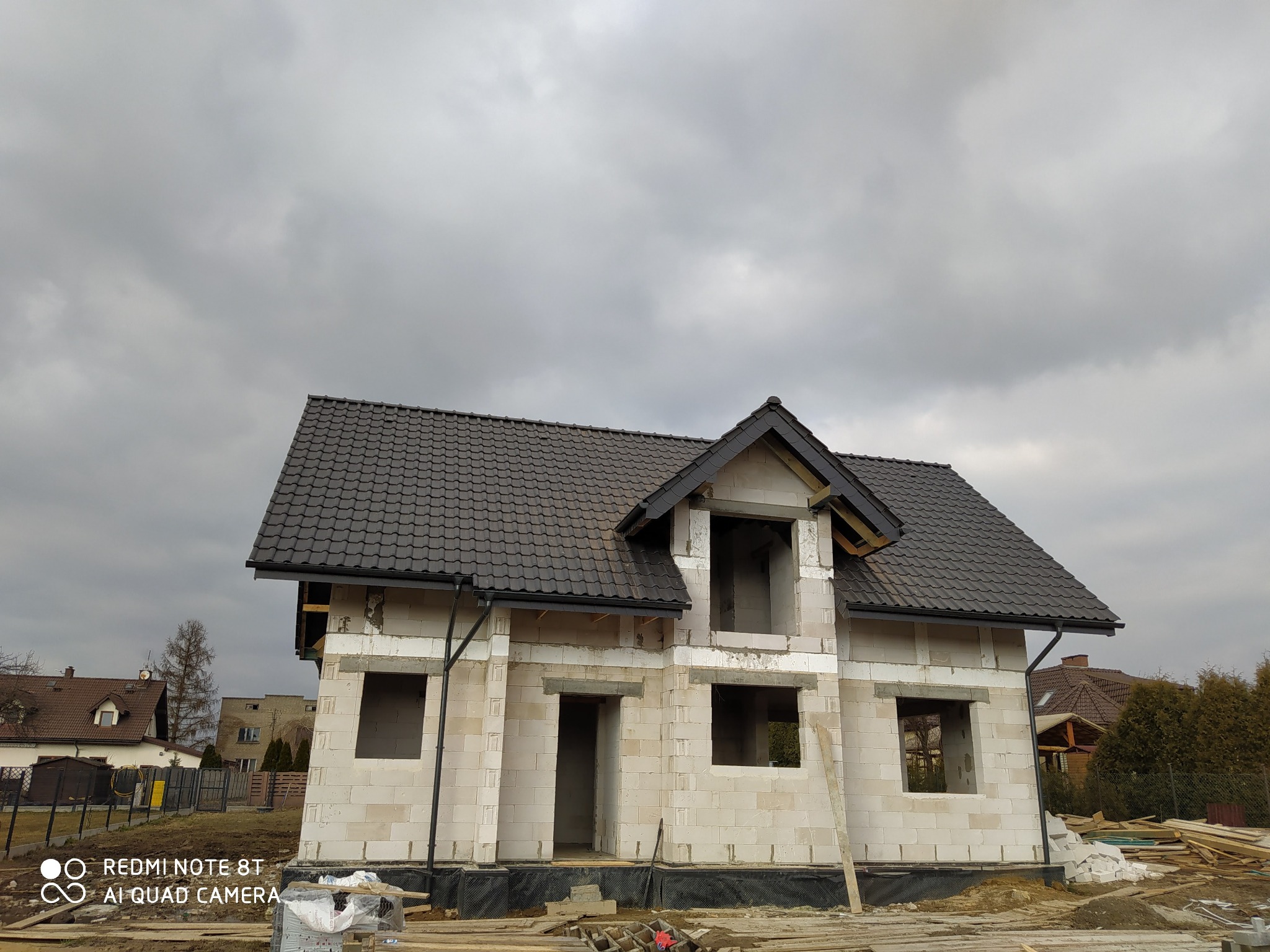 Surowy stan domu jednorodzinnego z białych bloczków, z ciemną dachówką, widoczne przygotowane drewno budowlane na pierwszym planie, pochmurne niebo w tle.