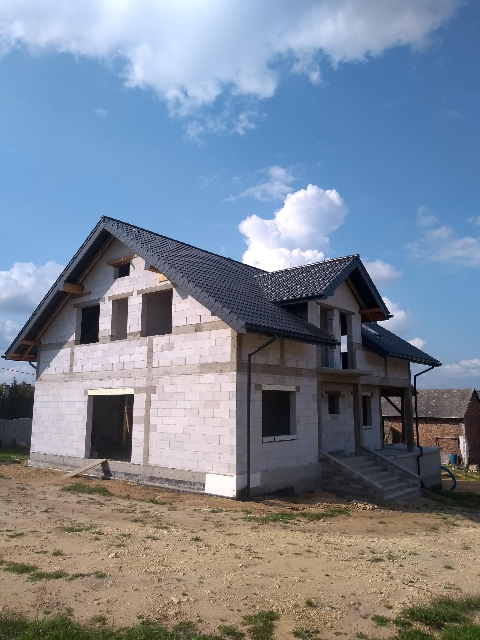 Dwukondygnacyjny dom w trakcie budowy, ściany z białych bloczków, dach z ciemnej dachówki, widoczne niezabudowane otwory okienne i drzwiowe, teren wokół domu niezagospodarowany, błękitne niebo...