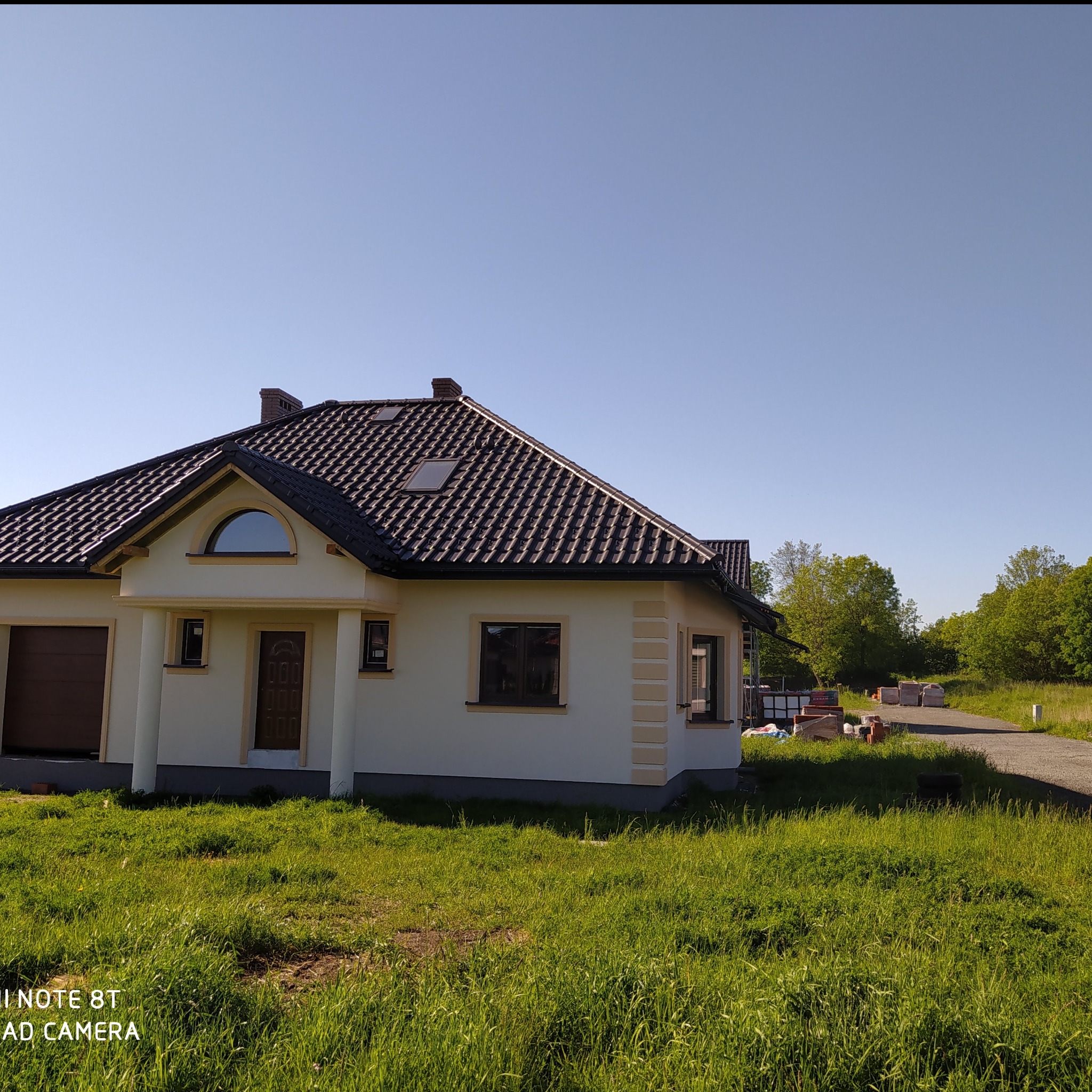 Gotowy dom jednorodzinny z brązową dachówką, kremową elewacją i elementami dekoracyjnymi, otoczony zielonym trawnikiem, z widocznymi materiałami budowlanymi w tle.