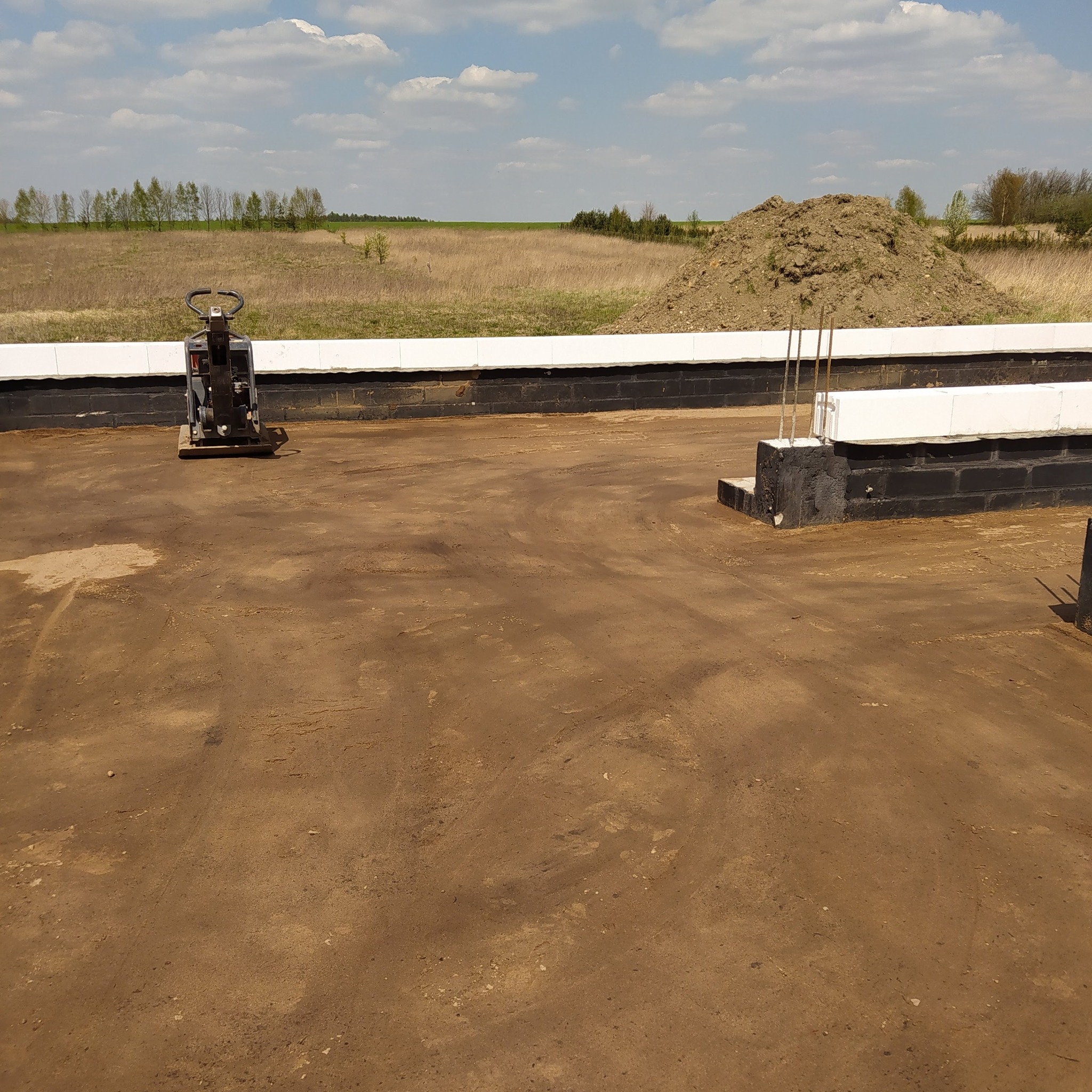 Zagęszczarka ubija piasek na wyrównanym terenie pod budowę fundamentów, widoczny fragment zbrojenia i murków z bloczków betonowych, w tle pole i niewielki kopiec ziemi.